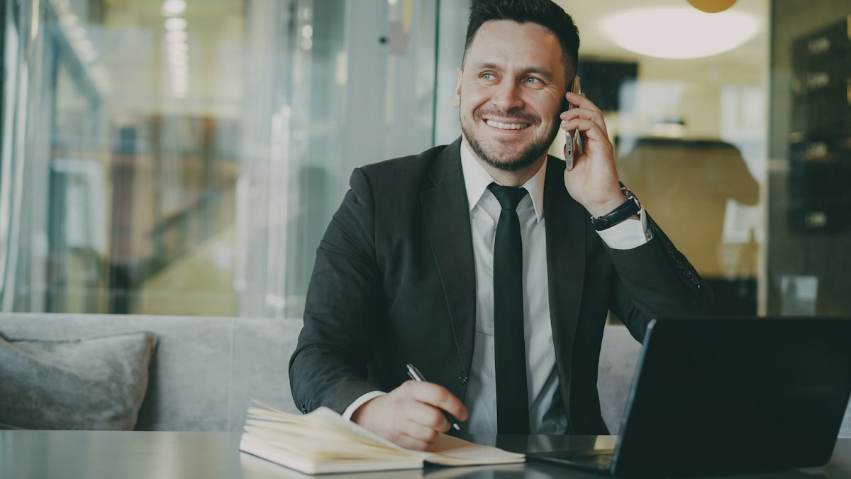 A salesman smiling on a phone call at his desk, taking notes